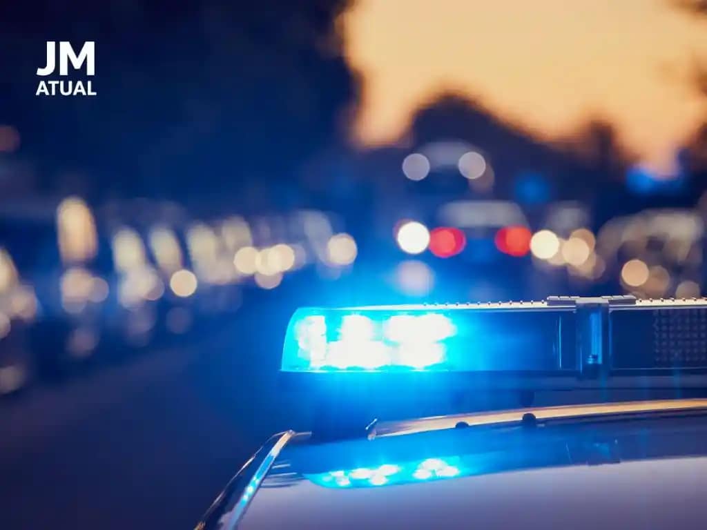 A imagem mostra uma viatura policial em destaque, com o giroflex azul aceso, ocupando a maior parte do enquadramento. O fundo está desfocado, com pontos de luz em tons avermelhados e amarelados, reforçando a ideia de ação policial noturna ou em ambiente urbano.