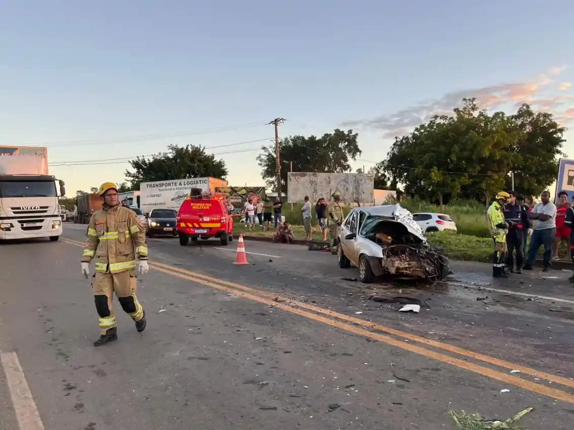 A imagem mostra a cena de um acidente na BR-381, com um carro de passeio fortemente danificado após uma colisão frontal. O veículo aparece com a parte dianteira completamente destruída e parado sobre a pista, cercado por destroços. No local, há atuação de equipes de resgate e segurança. Um bombeiro caminha pela rodovia, enquanto uma viatura do Corpo de Bombeiros está parada mais à frente, com o giroflex ligado. Cones foram posicionados para sinalizar o trecho e organizar o trânsito. Ao fundo, é possível ver caminhões, outros veículos parados e um grupo de pessoas observando a ocorrência às margens da rodovia. O cenário indica atendimento emergencial e interdição parcial da pista após o impacto.