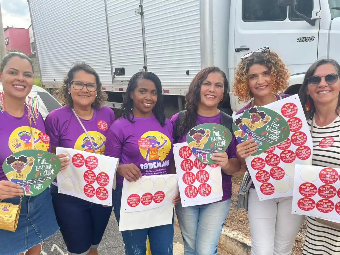 A imagem mostra seis mulheres reunidas ao ar livre, sorrindo para a câmera e segurando materiais de campanha. Elas vestem camisetas roxas com ilustrações e mensagens de conscientização sobre a proteção de crianças e adolescentes. Cada uma segura folhas com vários adesivos vermelhos e leques informativos com o slogan “Pule, brinque e cuide”, relacionados à campanha de combate à exploração sexual infantil.