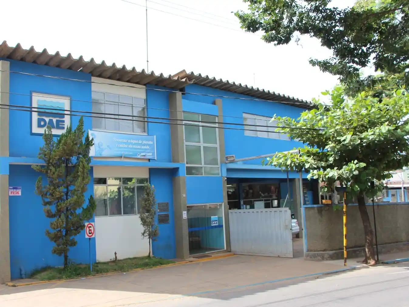 A imagem mostra a sede do Departamento de Águas e Esgotos (DAE) em um prédio de dois andares, pintado em tons de azul e branco. Na fachada, há o logotipo do DAE e uma placa com mensagem institucional sobre o uso consciente da água.