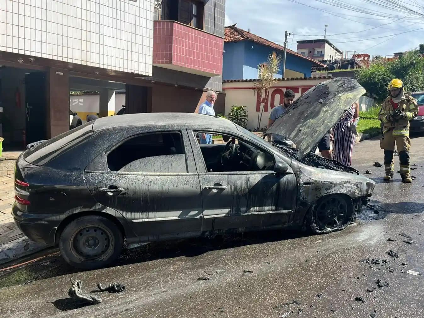 A imagem mostra um carro de passeio completamente queimado, parado ao lado do meio-fio em uma rua residencial durante o dia. O veículo apresenta a lataria escurecida e coberta por fuligem, com sinais claros de incêndio, especialmente na parte dianteira. O capô está aberto, expondo o compartimento do motor, que aparenta ter sido o ponto mais afetado pelas chamas. Nota-se, ainda, a presença de um militar do Corpo de Bombeiros de Monlevade.