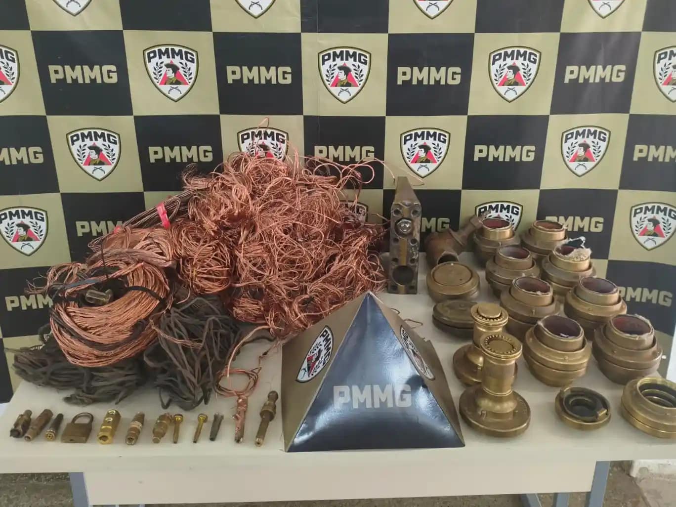 A imagem mostra diversos materiais apreendidos expostos sobre uma mesa, tendo ao fundo um painel institucional com a identificação da PMMG. Em destaque, há grande quantidade de fios e cabos de cobre enrolados e soltos, além de conexões metálicas, peças industriais de latão, registros, bicos e outros componentes utilizados em sistemas industriais. Os itens estão organizados lado a lado para registro da ocorrência, com um suporte triangular da corporação posicionado ao centro da mesa. A foto registra o material recuperado em ação da Polícia de Monlevade, evidenciando a apreensão dos objetos e a atuação policial no combate à receptação no município.