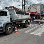 A imagem mostra uma operação tapa-buracos em via urbana, com equipes trabalhando na recuperação do asfalto. Em primeiro plano, há um caminhão basculante branco, estacionado parcialmente sobre a pista, sinalizado com cones de trânsito para organizar o fluxo de veículos. Ao fundo, trabalhadores uniformizados, alguns com coletes refletivos, realizam o reparo do pavimento, espalhando e nivelando o asfalto em um trecho da rua. A área está isolada para garantir a segurança durante o serviço.