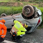 Bombeiros militares atuam no atendimento a um carro capotado na BR-262, em São Domingos do Prata. O veículo está virado de cabeça para baixo às margens da rodovia, enquanto os socorristas realizam procedimentos de segurança e avaliação no local, com vegetação ao redor da pista.