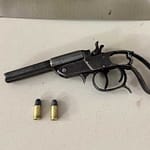 Imagem mostra uma arma de fogo do tipo garrucha apoiada sobre uma superfície clara, acompanhada de duas munições intactas posicionadas à frente. Ao fundo, aparece um suporte metálico em formato triangular com o emblema da Polícia Militar, indicando que o material foi apreendido e apresentado em ambiente institucional. A cena sugere registro oficial de apreensão realizada pelas forças de segurança.