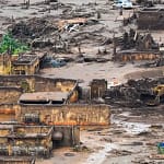 Imagem aérea mostra área devastada pelo rompimento da barragem em Brumadinho. Construções aparecem parcialmente soterradas pela lama, com paredes destruídas e estruturas expostas. O terreno está coberto por rejeitos de mineração, entulho e troncos de árvores.