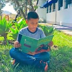 A imagem mostra uma criança sentada sobre a grama, em um ambiente externo e arborizado, lendo um livro infantil. Ela está sentada com as pernas cruzadas, concentrada na leitura, segurando um livro de capa verde com ilustrações coloridas.