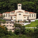 A imagem mostra a Igreja São José Operário em destaque, situada em uma área elevada e cercada por vegetação densa, com morros e árvores ao fundo. O templo possui arquitetura imponente e simétrica, com fachada clara, telhados em tom avermelhado e uma torre central que se eleva acima do conjunto principal. Na parte frontal, observa-se uma grande cruz fixada na fachada, elemento central da composição visual. As laterais apresentam janelas altas e arqueadas, reforçando o estilo religioso tradicional. O acesso à igreja é feito por escadarias e caminhos em níveis, acompanhando o relevo do terreno.