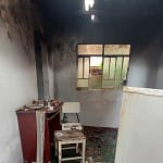 O interior de uma residência aparece severamente danificado por incêndio. As paredes e o teto estão cobertos por fuligem escura, com sinais evidentes de que o fogo se concentrou principalmente na parte superior do cômodo. Há uma abertura no telhado por onde entra luz natural. No ambiente, é possível ver móveis parcialmente queimados, como um pequeno armário, uma cadeira e uma mesa, além de objetos espalhados pelo chão.