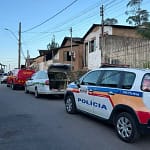 A foto mostra uma rua residencial com viaturas da Polícia Militar e do Corpo de Bombeiros estacionadas em fila ao longo da via. Em primeiro plano, aparece uma viatura da Polícia Militar de Minas Gerais, identificada com as cores branca, azul, vermelha e laranja. Logo à frente, há um veículo branco com o porta-malas aberto, seguido por uma viatura vermelha do Corpo de Bombeiros Militar.