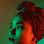 A imagem mostra o retrato de uma mulher vista de perfil, com o rosto levemente voltado para cima e o olhar direcionado para fora do enquadramento. Ela usa um turbante amarrado na cabeça, cobrindo totalmente o cabelo. A iluminação é artística e contrastante, com luz verde incidindo sobre parte do rosto e luz vermelha sobre a outra, criando um efeito dramático. O fundo é liso e desfocado, em tons suaves, destacando a expressão serena e contemplativa da mulher.