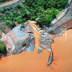 Imagem aérea mostra área afetada por rompimento de estrutura de contenção, com grande volume de lama alaranjada avançando por um vale, estradas danificadas, vegetação ao redor e trechos de terra expostos, evidenciando impacto ambiental e erosão do terreno.