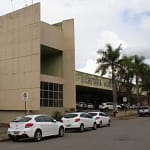 A foto mostra a fachada de um prédio público de grande porte, identificado como Secretaria Municipal de Saúde. O edifício tem arquitetura moderna, com estrutura em tons claros, linhas retas e um bloco vertical destacado na parte frontal. À frente do prédio, há uma via urbana com vários carros estacionados em fila ao longo do meio-fio.