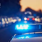 A imagem mostra uma viatura policial em destaque, com o giroflex azul aceso, ocupando a maior parte do enquadramento. O fundo está desfocado, com pontos de luz em tons avermelhados e amarelados, reforçando a ideia de ação policial noturna ou em ambiente urbano.