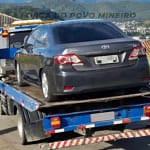 Um automóvel sedan de cor cinza escuro está sendo transportado sobre a plataforma de um caminhão-guincho azul, em via urbana. O veículo aparece visto pela traseira, com a placa parcialmente desfocada. Ao fundo, é possível observar casas, vegetação e uma área urbana em região montanhosa.