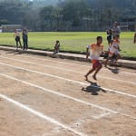 A imagem retrata uma prova de atletismo infantil, realizada em uma pista de terra demarcada, onde crianças disputam uma corrida em clima de competição e integração. Os atletas, usando camisetas padronizadas com numeração, correm descalços ou com calçados leves.