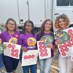 A imagem mostra seis mulheres reunidas ao ar livre, sorrindo para a câmera e segurando materiais de campanha. Elas vestem camisetas roxas com ilustrações e mensagens de conscientização sobre a proteção de crianças e adolescentes. Cada uma segura folhas com vários adesivos vermelhos e leques informativos com o slogan “Pule, brinque e cuide”, relacionados à campanha de combate à exploração sexual infantil.