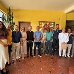 A foto mostra um grupo de onze pessoas reunidas em ambiente interno, após uma reunião institucional para tratar da preservação da Igreja São José Operário. Os participantes estão lado a lado, posando para o registro, com expressões sorridentes e postura informal, sugerindo clima de diálogo e cooperação. O espaço possui paredes amarelas, piso em cerâmica avermelhada e iluminação natural vinda de uma janela lateral. Ao fundo, há um quadro artístico colorido na parede e uma planta de grande porte próxima à janela.