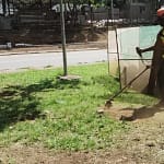 A imagem mostra um trabalhador realizando serviço de roçagem em uma área verde urbana, utilizando uma roçadeira manual motorizada. Ele veste equipamentos de proteção individual, como capacete com viseira, luvas, botas e uniforme de trabalho, indicando atividade de manutenção urbana.