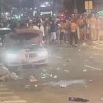 A imagem mostra uma avenida à noite, com grande concentração de pessoas ocupando a pista, com a presença da polícia em Monlevade. No primeiro plano, um carro branco aparece parado com os faróis acesos, cercado por lixo espalhado pelo asfalto, como copos e garrafas descartáveis. Ao fundo, é possível ver uma multidão reunida em frente aos veículos, ocupando os dois sentidos da via. A iluminação é típica de rua, com postes e luzes de estabelecimentos ao redor.