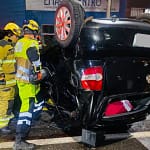 A imagem mostra um veículo preto capotado após acidente em Monlevade, com as rodas voltadas para cima, imobilizado no meio de uma via urbana durante a noite. O automóvel apresenta danos significativos na lataria, especialmente na parte traseira e lateral, indicando a força do impacto. Ao lado do carro, dois militares do Corpo de Bombeiros, devidamente equipados com capacetes, lanternas e uniformes de proteção, atuam no atendimento à ocorrência.