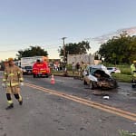 A imagem mostra a cena de um acidente na BR-381, com um carro de passeio fortemente danificado após uma colisão frontal. O veículo aparece com a parte dianteira completamente destruída e parado sobre a pista, cercado por destroços. No local, há atuação de equipes de resgate e segurança. Um bombeiro caminha pela rodovia, enquanto uma viatura do Corpo de Bombeiros está parada mais à frente, com o giroflex ligado. Cones foram posicionados para sinalizar o trecho e organizar o trânsito. Ao fundo, é possível ver caminhões, outros veículos parados e um grupo de pessoas observando a ocorrência às margens da rodovia. O cenário indica atendimento emergencial e interdição parcial da pista após o impacto.