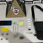 A imagem mostra os materiais apreendidos pela Polícia Militar, organizados sobre uma mesa, tendo ao fundo um painel com a identificação da corporação e um suporte com o brasão da PMMG. Entre os itens expostos estão notas e moedas em dinheiro, um aparelho celular, um cartão bancário, um frasco contendo lança-perfume, comprimidos de ecstasy, papelotes de substância semelhante à cocaína, uma porção de maconha, um cigarro artesanal e uma bolsa preta. O material foi apresentado como resultado de uma ocorrência registrada em Bela Vista de Minas.