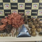 A imagem mostra diversos materiais apreendidos expostos sobre uma mesa, tendo ao fundo um painel institucional com a identificação da PMMG. Em destaque, há grande quantidade de fios e cabos de cobre enrolados e soltos, além de conexões metálicas, peças industriais de latão, registros, bicos e outros componentes utilizados em sistemas industriais. Os itens estão organizados lado a lado para registro da ocorrência, com um suporte triangular da corporação posicionado ao centro da mesa. A foto registra o material recuperado em ação da Polícia de Monlevade, evidenciando a apreensão dos objetos e a atuação policial no combate à receptação no município.