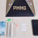 A imagem mostra materiais apreendidos pela Polícia Militar, dispostos sobre uma superfície clara, com um suporte ao fundo exibindo a sigla “PMMG”. Entre os itens estão uma cédula de R$ 100, um frasco plástico contendo lança-perfume, três papelotes de substância semelhante à cocaína, um comprimido de ecstasy e um aparelho celular. Os objetos estão organizados lado a lado, compondo o registro da ocorrência policial. Notícias de nova era