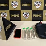 A imagem mostra os materiais apreendidos pela Polícia Militar durante a ocorrência registrada em Nova Era. Sobre uma mesa branca, estão expostos três frascos de lança-perfume, um aparelho celular com capa vermelha e uma bolsa preta tipo pochete. Ao lado dos objetos, aparece um suporte triangular com a sigla da corporação, posicionado diante de um painel ao fundo com o padrão xadrez nas cores preta e amarela, estampado com o emblema da PMMG. A foto integra as notícias de Nova Era relacionadas à prisão de dois suspeitos por tráfico de drogas durante evento carnavalesco no Centro da cidade.