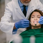 A imagem mostra um atendimento odontológico em ambiente clínico. Um profissional da odontologia, vestindo jaleco branco e luvas azuis, realiza um procedimento na boca de uma paciente sentada na cadeira odontológica. O dentista utiliza instrumentos próprios da área, como espelho e sonda, enquanto a paciente permanece reclinada, com expressão tranquila e sorridente.