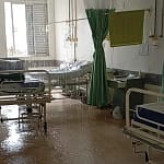 Hospital em João Monlevade com enfermaria masculina parcialmente alagada após vazamento no teto. A imagem mostra leitos hospitalares posicionados lado a lado, com água escorrendo do forro e formando poças no piso. Parte do teto apresenta infiltração visível, e o chão está molhado em diversos pontos, refletindo a iluminação do ambiente.