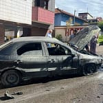 A imagem mostra um carro de passeio completamente queimado, parado ao lado do meio-fio em uma rua residencial durante o dia. O veículo apresenta a lataria escurecida e coberta por fuligem, com sinais claros de incêndio, especialmente na parte dianteira. O capô está aberto, expondo o compartimento do motor, que aparenta ter sido o ponto mais afetado pelas chamas. Nota-se, ainda, a presença de um militar do Corpo de Bombeiros de Monlevade.