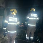 A imagem mostra bombeiros em João Monlevade atuando durante a noite em uma ocorrência. Dois militares do Corpo de Bombeiros aparecem de costas, utilizando capacetes amarelos e uniformes com faixas refletivas, que brilham sob a iluminação do local. O ambiente está escuro e com presença de fumaça, sugerindo atendimento a um incêndio recente. No chão, é possível observar equipamentos e materiais próximos a uma área aparentemente queimada. A cena transmite a atuação da equipe no trabalho de rescaldo e verificação da área atingida.