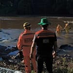 Em Nova Era, equipes do Corpo de Bombeiros e da polícia acompanham uma operação de resgate em um trecho do Rio Piracicaba. Na imagem, militares uniformizados estão próximos às margens e dentro da água, entre rochas, realizando o trabalho de recuperação do corpo.