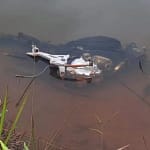 A imagem mostra uma motocicleta parcialmente submersa em uma lagoa, com a água de coloração turva e barrenta. Apenas parte da estrutura metálica do veículo permanece visível na superfície, destacando-se o motor exposto e componentes do chassi. A motocicleta aparenta estar sem rodas, carenagem e outras peças, indicando que foi desmontada ou “depenada” antes de ser abandonada no local.