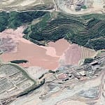Imagem aérea mostra parte do Complexo Minerário de Itabira, com destaque para áreas de lavra, estruturas de beneficiamento e uma grande bacia com rejeitos de coloração avermelhada, cercada por taludes em formato de degraus. Ao redor, é possível observar vegetação, vias internas de acesso e instalações industriais ligadas à atividade mineradora. Registro relacionado às notícias de Itabira sobre projetos e operações no setor mineral.