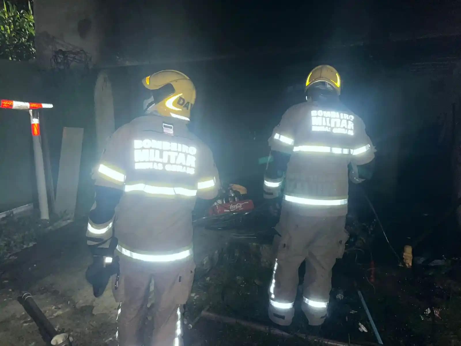 A imagem mostra bombeiros em João Monlevade atuando durante a noite em uma ocorrência. Dois militares do Corpo de Bombeiros aparecem de costas, utilizando capacetes amarelos e uniformes com faixas refletivas, que brilham sob a iluminação do local. O ambiente está escuro e com presença de fumaça, sugerindo atendimento a um incêndio recente. No chão, é possível observar equipamentos e materiais próximos a uma área aparentemente queimada. A cena transmite a atuação da equipe no trabalho de rescaldo e verificação da área atingida.