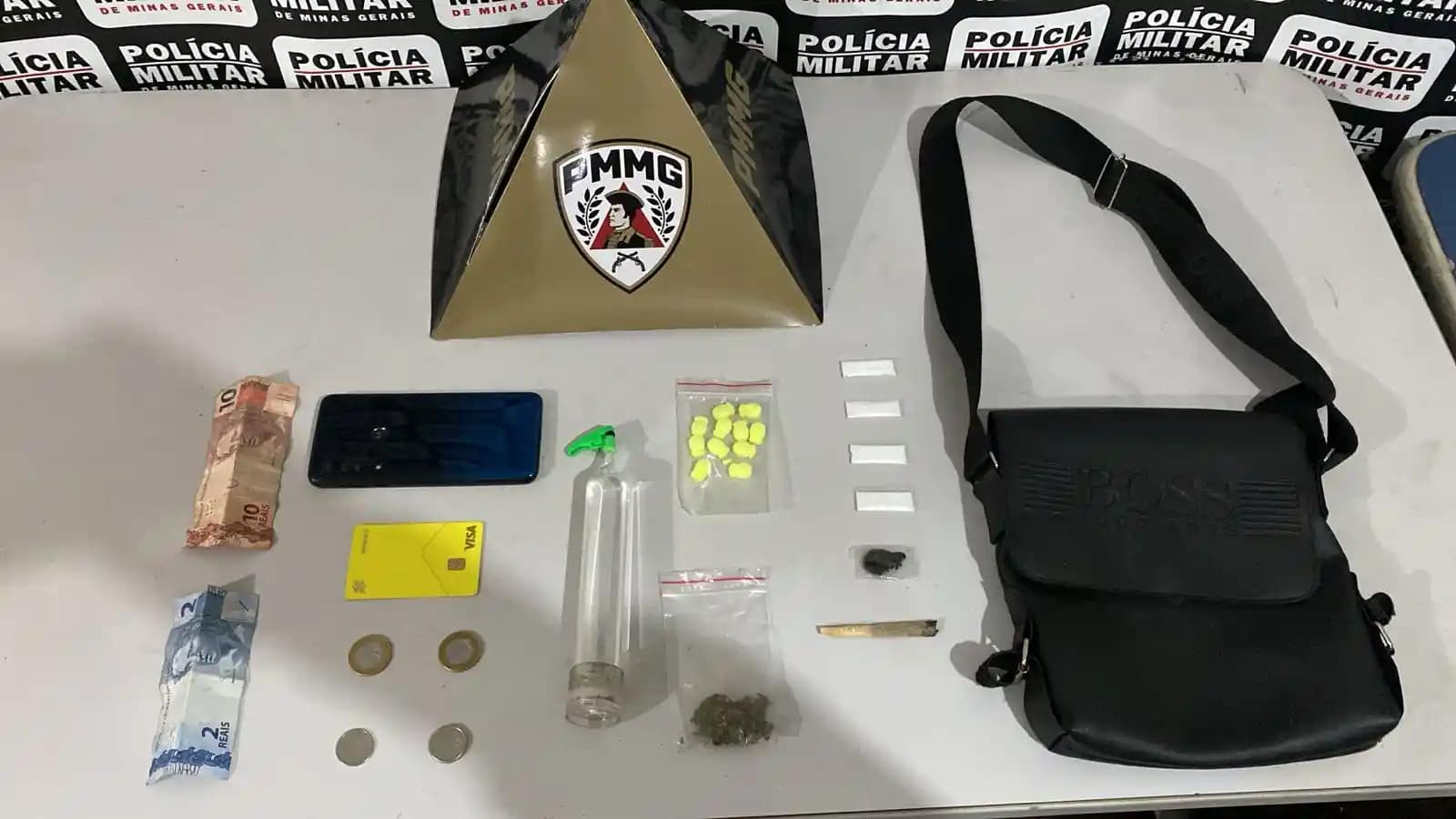A imagem mostra os materiais apreendidos pela Polícia Militar, organizados sobre uma mesa, tendo ao fundo um painel com a identificação da corporação e um suporte com o brasão da PMMG. Entre os itens expostos estão notas e moedas em dinheiro, um aparelho celular, um cartão bancário, um frasco contendo lança-perfume, comprimidos de ecstasy, papelotes de substância semelhante à cocaína, uma porção de maconha, um cigarro artesanal e uma bolsa preta. O material foi apresentado como resultado de uma ocorrência registrada em Bela Vista de Minas.