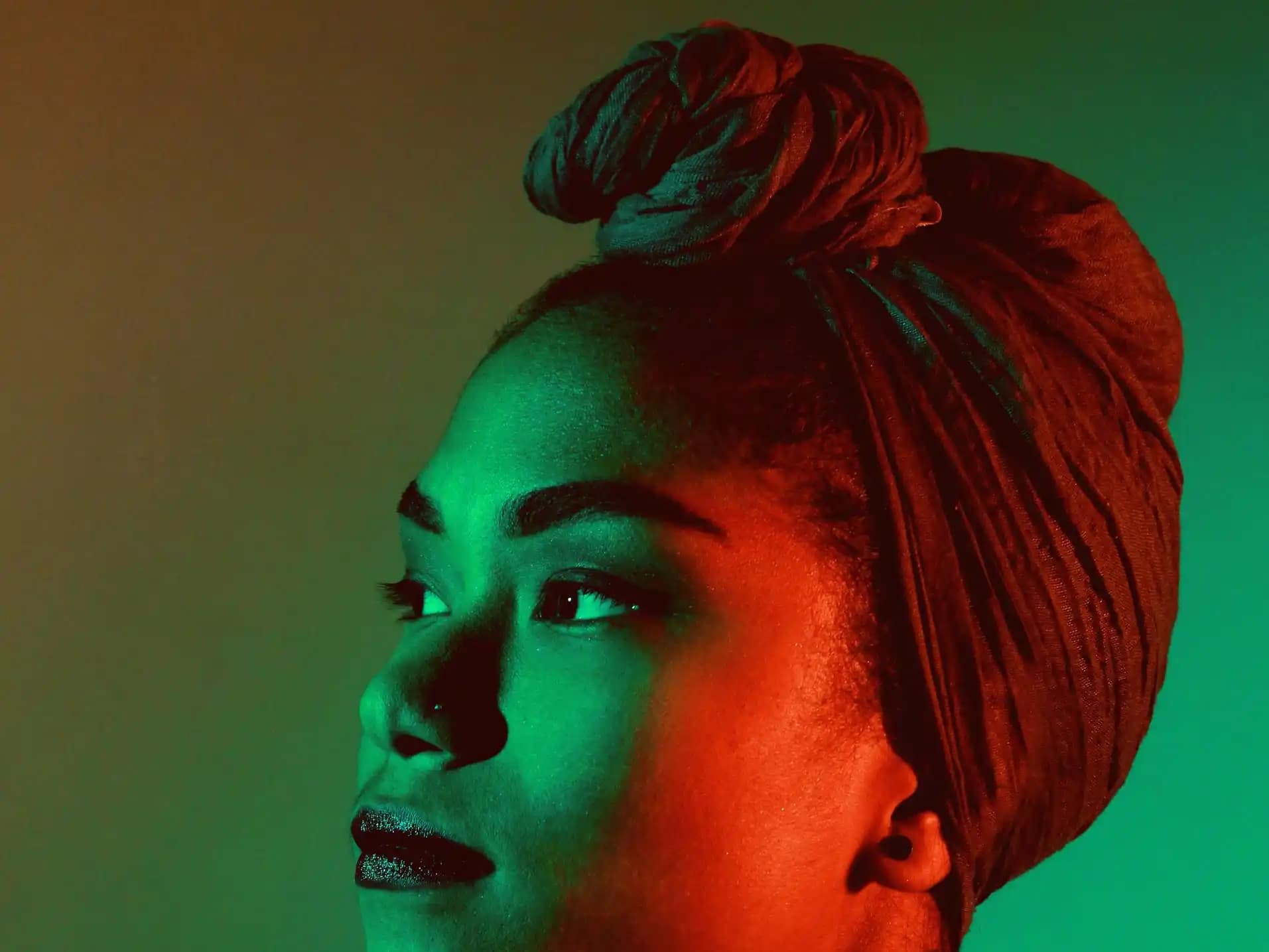 A imagem mostra o retrato de uma mulher vista de perfil, com o rosto levemente voltado para cima e o olhar direcionado para fora do enquadramento. Ela usa um turbante amarrado na cabeça, cobrindo totalmente o cabelo. A iluminação é artística e contrastante, com luz verde incidindo sobre parte do rosto e luz vermelha sobre a outra, criando um efeito dramático. O fundo é liso e desfocado, em tons suaves, destacando a expressão serena e contemplativa da mulher.