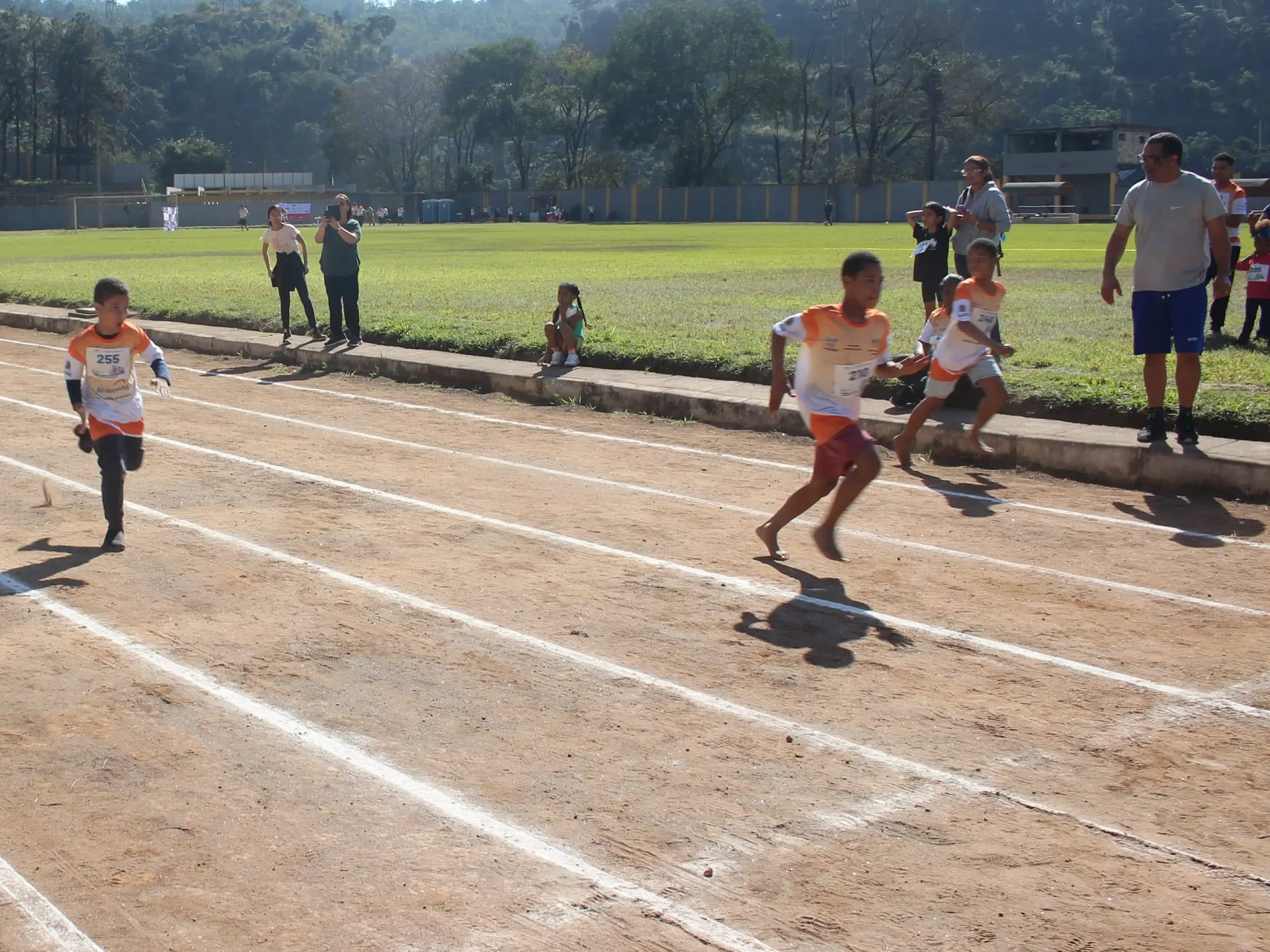 A imagem retrata uma prova de atletismo infantil, realizada em uma pista de terra demarcada, onde crianças disputam uma corrida em clima de competição e integração. Os atletas, usando camisetas padronizadas com numeração, correm descalços ou com calçados leves.