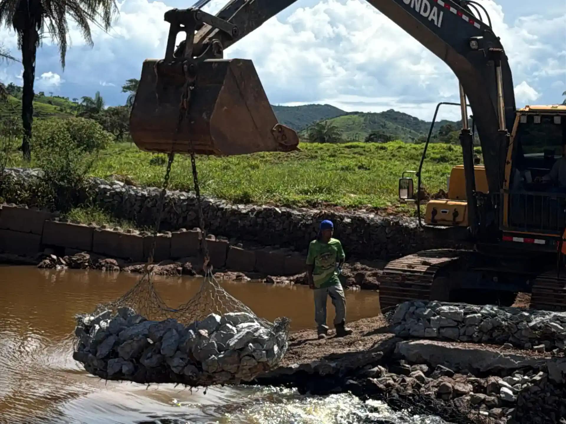 A imagem mostra uma obra de reforço em um curso d’água, em uma barragem ou dique. Uma escavadeira hidráulica, posicionada à margem do rio, utiliza a concha para içar uma rede metálica cheia de pedras, conhecida como gabião, que será utilizada na contenção da estrutura. Ao lado da máquina, um trabalhador acompanha a operação em pé sobre o terreno de terra e pedras.