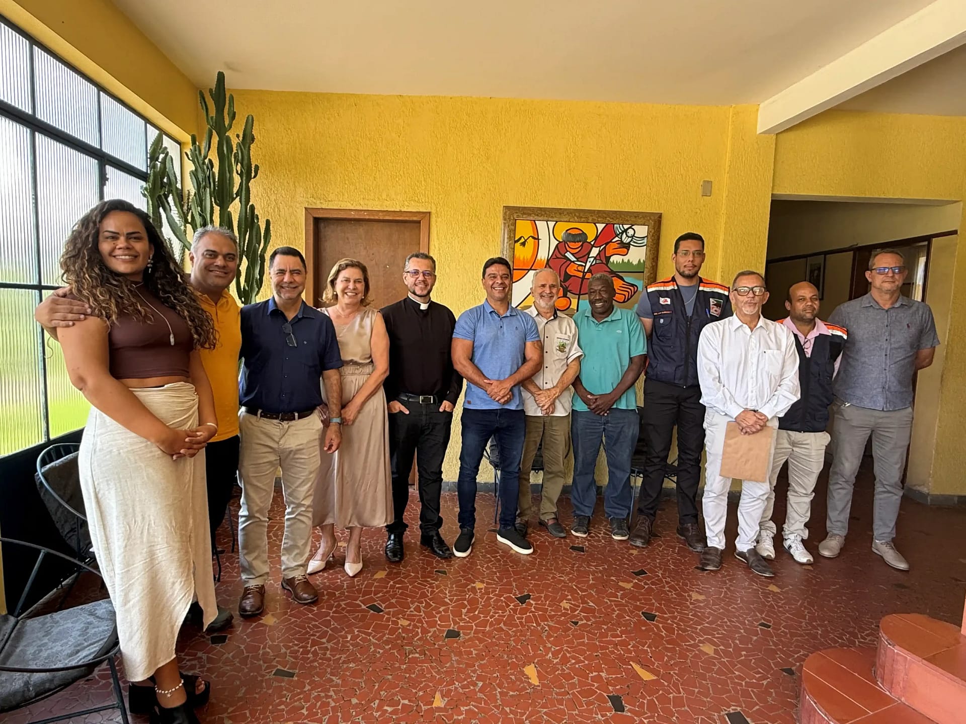 A foto mostra um grupo de onze pessoas reunidas em ambiente interno, após uma reunião institucional para tratar da preservação da Igreja São José Operário. Os participantes estão lado a lado, posando para o registro, com expressões sorridentes e postura informal, sugerindo clima de diálogo e cooperação. O espaço possui paredes amarelas, piso em cerâmica avermelhada e iluminação natural vinda de uma janela lateral. Ao fundo, há um quadro artístico colorido na parede e uma planta de grande porte próxima à janela.