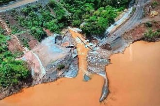 Imagem aérea mostra área afetada por rompimento de estrutura de contenção, com grande volume de lama alaranjada avançando por um vale, estradas danificadas, vegetação ao redor e trechos de terra expostos, evidenciando impacto ambiental e erosão do terreno.