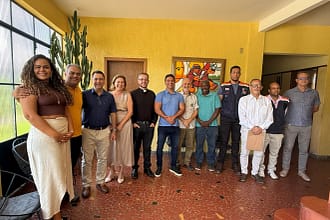 A foto mostra um grupo de onze pessoas reunidas em ambiente interno, após uma reunião institucional para tratar da preservação da Igreja São José Operário. Os participantes estão lado a lado, posando para o registro, com expressões sorridentes e postura informal, sugerindo clima de diálogo e cooperação. O espaço possui paredes amarelas, piso em cerâmica avermelhada e iluminação natural vinda de uma janela lateral. Ao fundo, há um quadro artístico colorido na parede e uma planta de grande porte próxima à janela.