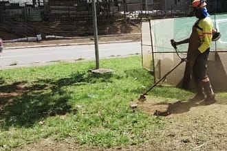A imagem mostra um trabalhador realizando serviço de roçagem em uma área verde urbana, utilizando uma roçadeira manual motorizada. Ele veste equipamentos de proteção individual, como capacete com viseira, luvas, botas e uniforme de trabalho, indicando atividade de manutenção urbana.