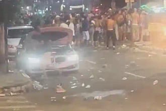 A imagem mostra uma avenida à noite, com grande concentração de pessoas ocupando a pista, com a presença da polícia em Monlevade. No primeiro plano, um carro branco aparece parado com os faróis acesos, cercado por lixo espalhado pelo asfalto, como copos e garrafas descartáveis. Ao fundo, é possível ver uma multidão reunida em frente aos veículos, ocupando os dois sentidos da via. A iluminação é típica de rua, com postes e luzes de estabelecimentos ao redor.