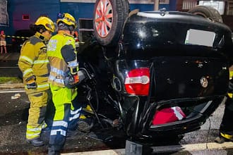 A imagem mostra um veículo preto capotado após acidente em Monlevade, com as rodas voltadas para cima, imobilizado no meio de uma via urbana durante a noite. O automóvel apresenta danos significativos na lataria, especialmente na parte traseira e lateral, indicando a força do impacto. Ao lado do carro, dois militares do Corpo de Bombeiros, devidamente equipados com capacetes, lanternas e uniformes de proteção, atuam no atendimento à ocorrência.