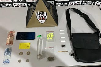A imagem mostra os materiais apreendidos pela Polícia Militar, organizados sobre uma mesa, tendo ao fundo um painel com a identificação da corporação e um suporte com o brasão da PMMG. Entre os itens expostos estão notas e moedas em dinheiro, um aparelho celular, um cartão bancário, um frasco contendo lança-perfume, comprimidos de ecstasy, papelotes de substância semelhante à cocaína, uma porção de maconha, um cigarro artesanal e uma bolsa preta. O material foi apresentado como resultado de uma ocorrência registrada em Bela Vista de Minas.