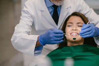 A imagem mostra um atendimento odontológico em ambiente clínico. Um profissional da odontologia, vestindo jaleco branco e luvas azuis, realiza um procedimento na boca de uma paciente sentada na cadeira odontológica. O dentista utiliza instrumentos próprios da área, como espelho e sonda, enquanto a paciente permanece reclinada, com expressão tranquila e sorridente.